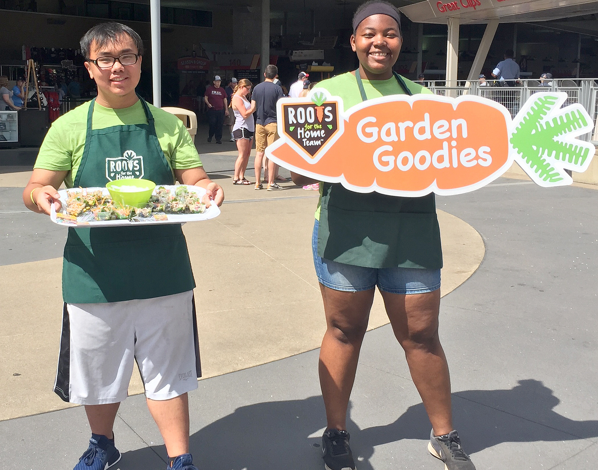 Two members of Roots for the Home Team, one holds up a sign that says, "Roots for the Home Team, Garden Goodies."